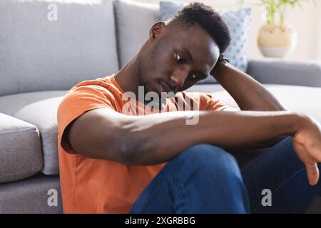 Thoughtful african american man sitting on floor in sunny living room holding head. Mental health, depression, loneliness, emotion, domestic life and Stock Photo