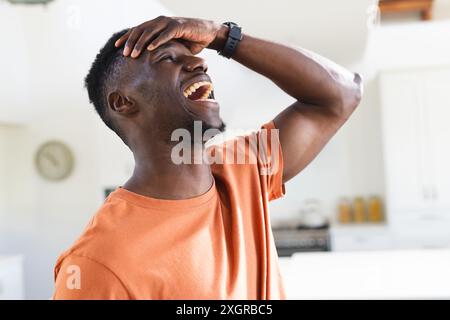 Happy african american man in sunny kitchen, holding head and laughing. Wellbeing, relaxation, domestic life and lifestyle, unaltered. Stock Photo