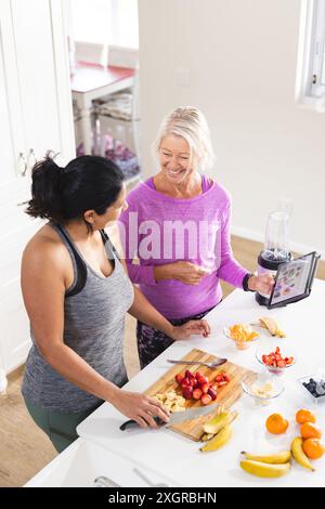 Happy diverse senior female friends using tablet preparing fruit for smoothie in kitchen, copy space. Communication, food, nutrition, friendship, dome Stock Photo