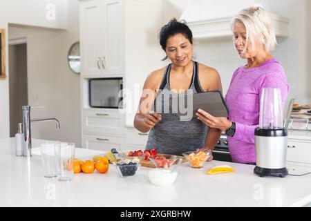 Happy diverse senior female friends using tablet preparing fruit smoothie in kitchen, copy space. Communication, food, nutrition, friendship, domestic Stock Photo