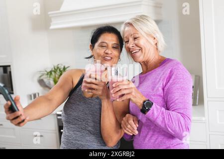 Happy diverse senior female friends drinking fruit smoothie taking selfie in kitchen. Communication, food, nutrition, friendship, domestic life and he Stock Photo