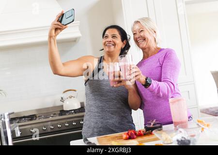 Happy diverse senior female friends drinking fruit smoothie in kitchen taking selfie. Communication, food, nutrition, friendship, domestic life and he Stock Photo