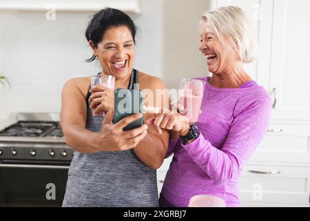 Happy diverse senior female friends drinking smoothies in kitchen, looking at smartphone laughing. Food, nutrition, friendship, domestic life and heal Stock Photo