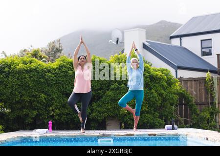 Two diverse senior female friends practicing yoga standing stretching in sunny garden. Yoga, wellbeing, exercise, friendship and healthy senior lifest Stock Photo