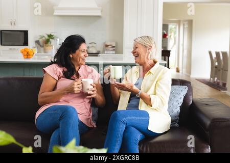 Two happy diverse senior female friends having coffee and talking on sofa in living room. Friendship, relaxation, retirement, domestic life and senior Stock Photo