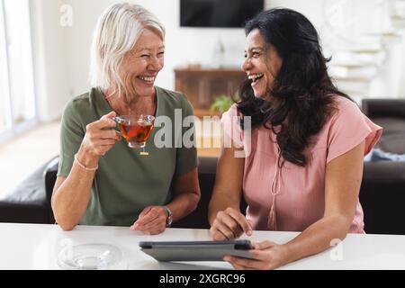 Two happy diverse senior female friends having tea, using tablet and laughing in kitchen. Friendship, communication, relaxation, retirement, domestic Stock Photo