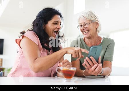 Two happy diverse senior female friends using smartphone and talking in kitchen. Friendship, communication, relaxation, retirement, domestic life and Stock Photo