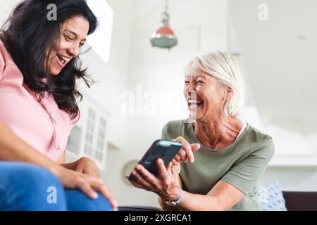 Two happy diverse senior female friends using smartphone, laughing in living room. Friendship, communication, technology, retirement, domestic life an Stock Photo