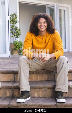Young biracial man sitting on steps outside a home. He exudes a relaxed and welcoming demeanor on a sunny day, unaltered. Stock Photo