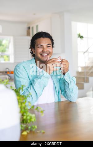 Young biracial gay couple enjoys warm drinks at home, with copy space. They're smiling and relaxed in a bright, modern kitchen setting. Stock Photo