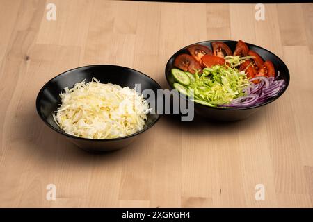 The two black bowls with fresh vegetable salad, one with shredded cabbage and the other with tomatoes on wooden table Stock Photo