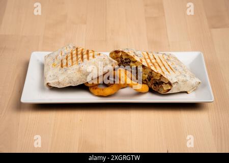 A grilled wrap filled with chicken and vegetables served with onion rings on a white plate over wooden table Stock Photo