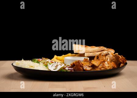 A delicious plate of mixed salad, fries, pita bread, chicken and sauce on a wooden table against black background Stock Photo