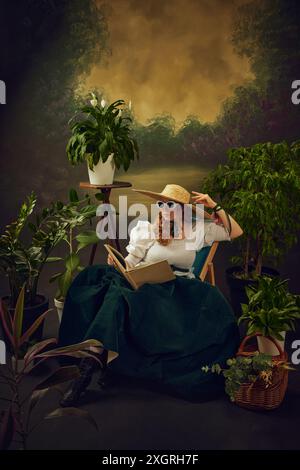 Portrait of elegant young woman in retro style glasses, straw hat, and velvet skirt, sitting on chair around lush green plants, and reading book Stock Photo