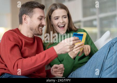 Happy overjoyed married couple using phone for online shopping smiling paying by credit card at home Stock Photo