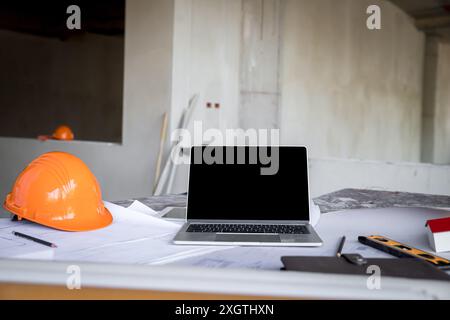 Laptop with helmet, blueprint and architects house model on desk in construction site. Stock Photo
