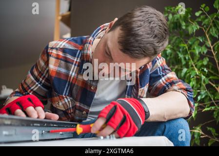 Man in plaid shirt and gloves focuses on repairing an electronic device using screwdriver, showcasing DIY skills.  Stock Photo