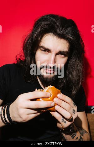 A young man with long, dark hair savors a delicious burger against a bright red background, embodying a relaxed and inviting barbecue theme Stock Photo