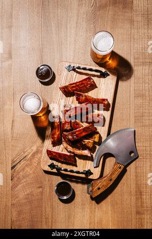 A rustic barbecue setting with juicy, grilled ribs laid out on a wooden cutting board, accompanied by two glasses of frothy beer and a unique cleaver Stock Photo