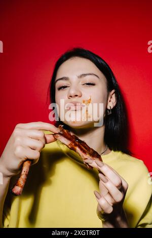 A young woman enjoys eating barbecue ribs, with sauce smeared on her face, set against a vibrant red backdrop, highlighting a joyful and messy dining Stock Photo