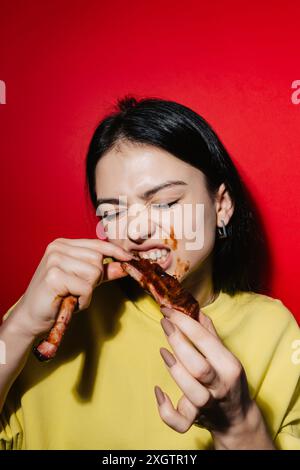 A vibrant image featuring a young woman in a yellow shirt, joyfully eating barbecue ribs Her hands and cheeks are playfully smeared with sauce, set ag Stock Photo