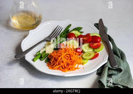 Healthy vegetable salad featuring asparagus with cheese, halved cherry tomatoes, cucumber slices, and grated carrots on a white plate with dressing an Stock Photo