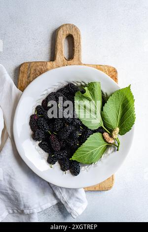 Top view of a white plate full of juicy, dark mulberries garnished with fresh green leaves, presented on a rustic wooden board. The image is composed Stock Photo