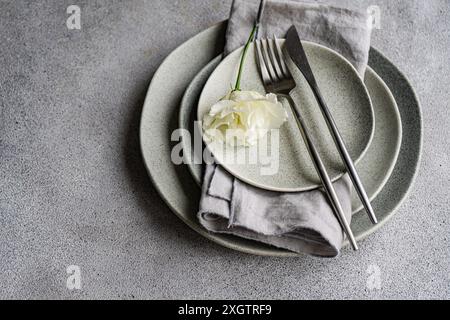 A tasteful table setting featuring stacks of gray ceramic plates, a silver fork and knife, and a delicate white Godetia flower, presented on a texture Stock Photo