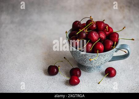 From above blue ornate ceramic bowl filled with fresh red cherries, situated on a textured grey surface. Several cherries are scattered around the bow Stock Photo