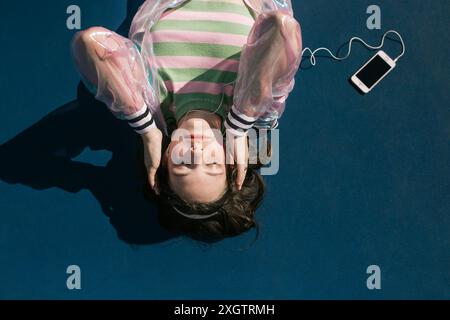 A young woman lies back on a blue tennis court, eyes closed and headphones on, enjoying music from a white smartphone lying beside her Stock Photo