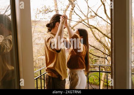 Side view of young man and woman share a playful moment dancing on a balcony amidst a backdrop of lush greenery. Stock Photo