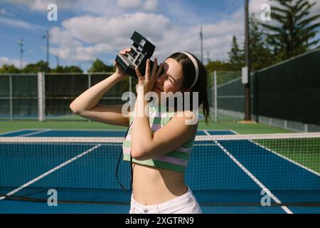 A stylish woman in a pastel-striped crop top and white pants takes photos with a vintage camera on a sunny tennis court The scene blends nostalgia wit Stock Photo