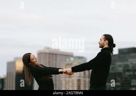 Side view of man and a woman dance on a city rooftop, holding hands, with a backdrop of urban high-rises. Stock Photo