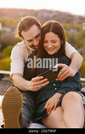 A young man and a woman share a gentle intimate moment, smiling while engaging in a smartphone on a rooftop during sunset. They are looking down at th Stock Photo