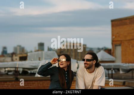 A man and a woman, both wearing sunglasses, share a joyful moment on a rooftop overlooking an urban skyline. The woman adjusts her sunglasses as both Stock Photo