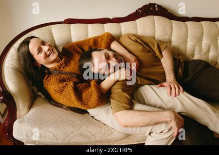 A happy woman and man sharing a playful moment as they lie on an ornate vintage sofa. The woman is looking at the camera with a joyful expression, whi Stock Photo