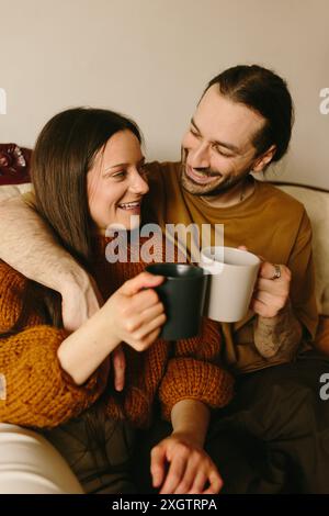 A Caucasian woman and man sitting closely on a cream-colored sofa, sharing a tender gaze. The woman, holding the man's hand, lovingly looks at him whi Stock Photo