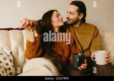 A Caucasian woman and man sitting closely on a cream-colored sofa, sharing a tender gaze. The woman, holding the man's hand, lovingly looks at him whi Stock Photo