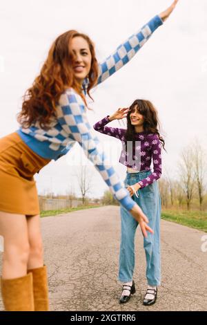 Two young women are captured in a playful moment on a deserted country road. One extends her hand as if dancing, while the other, sporting a casual sm Stock Photo