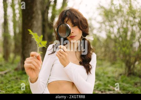 A young woman in a white dress enthusiastically examines a young plant using a magnifying glass in a lush woodland setting, displaying curiosity and e Stock Photo
