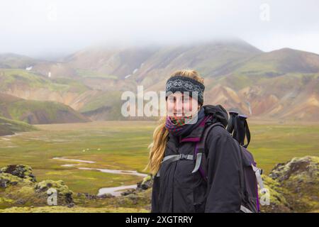A cheerful female hiker with a backpack smiles against a backdrop of the colorful mountainous landscapes of Iceland, featuring lush green valleys and Stock Photo