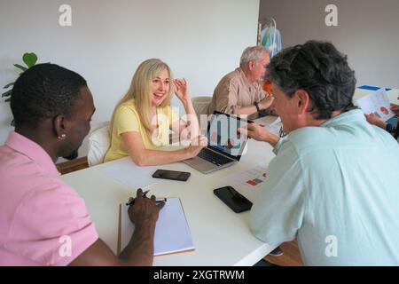 A multiethnic team engages in a business meeting around a white table, discussing data charts on a laptop in a well-lit room Stock Photo