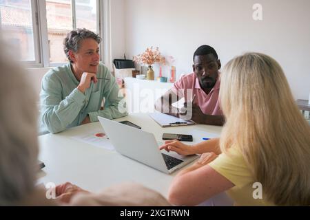 A diverse group of professionals engaged in a discussion at a white office table, focusing closely on a laptop screen and documents The room is bright Stock Photo