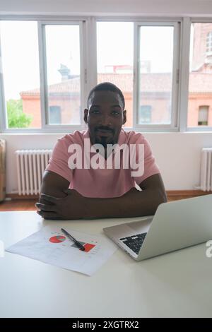 A black man sits at a white desk in a well-lit office space, smiling at the camera with a laptop and financial documents in front of him Stock Photo
