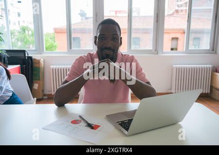 A confident African American man sits at his office desk, interacting with a laptop and business documents, projecting professionalism and focus in a Stock Photo