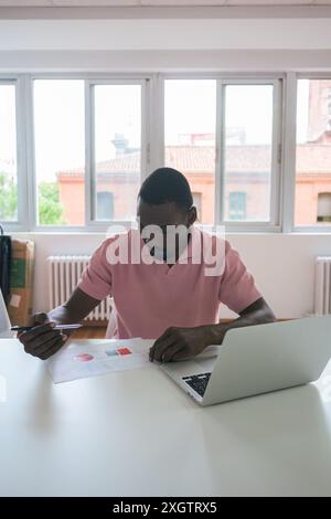 A focused black man reviews financial reports and uses a laptop at a well-lit office, suggesting productivity and business analysis in a contemporary Stock Photo