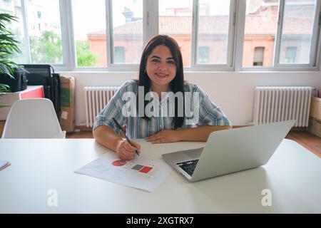 A cheerful young woman in a blue striped shirt sits at a bright office desk, working on a laptop while reviewing documents with a pen in hand Stock Photo