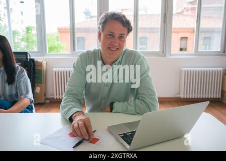 A mature businessman focuses on paperwork while sitting at a white table with a laptop, in a well-lit office environment, showcasing a professional an Stock Photo