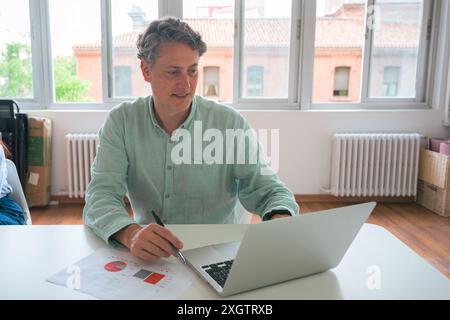 A mature professional male evaluates data on a laptop, jotting notes in a sunlit office, showcasing a typical business scene Stock Photo