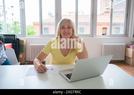 A cheerful mature woman smiles at the camera while working with a laptop in a well-lit office environment, demonstrating professionalism and engagemen Stock Photo
